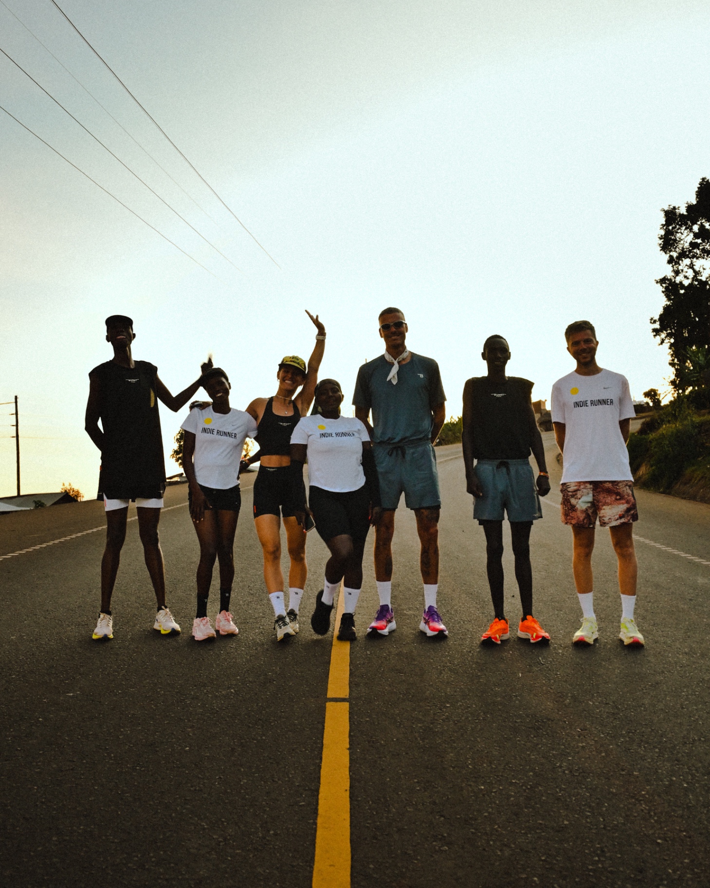 Seven runners standing side-by-side across the centre line of a rural road at sunrise